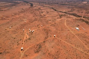 White dish shaped radio telescopes nad a large white building from the air on a red Australian desert backdrop. 