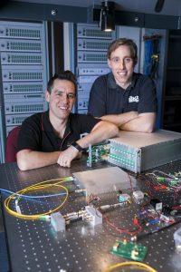 Sascha Schediwy and David Gozzard shown with an SKA-mid Transmitter Module and Sub-Rack enclosure. In the foreground is the Receiver Module to be installed at each antenna, and the background shows a mock-up of the entire 197-element system to be installed in the SKA’s central processor building. Credit: ICRAR.