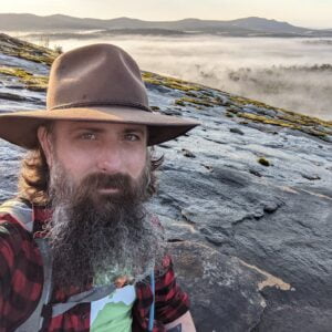 Photo of Tim Young in a hat sitting on a cliff overlooking a misty forest.