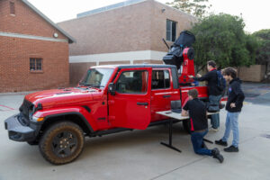 Three students working at the back of a red ute, with a telescope mounted in the tray pointed at the sky.