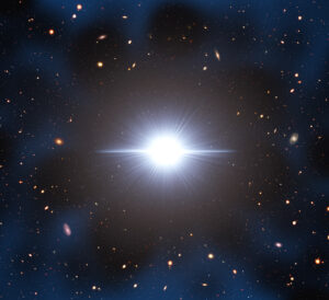 Artist Illustration of Early-Universe Quasar Cosmic Neighbourhood. Image shows a large, bright white star-like object in the middle of a dark sky, surrounded by many, much smaller galaxies.